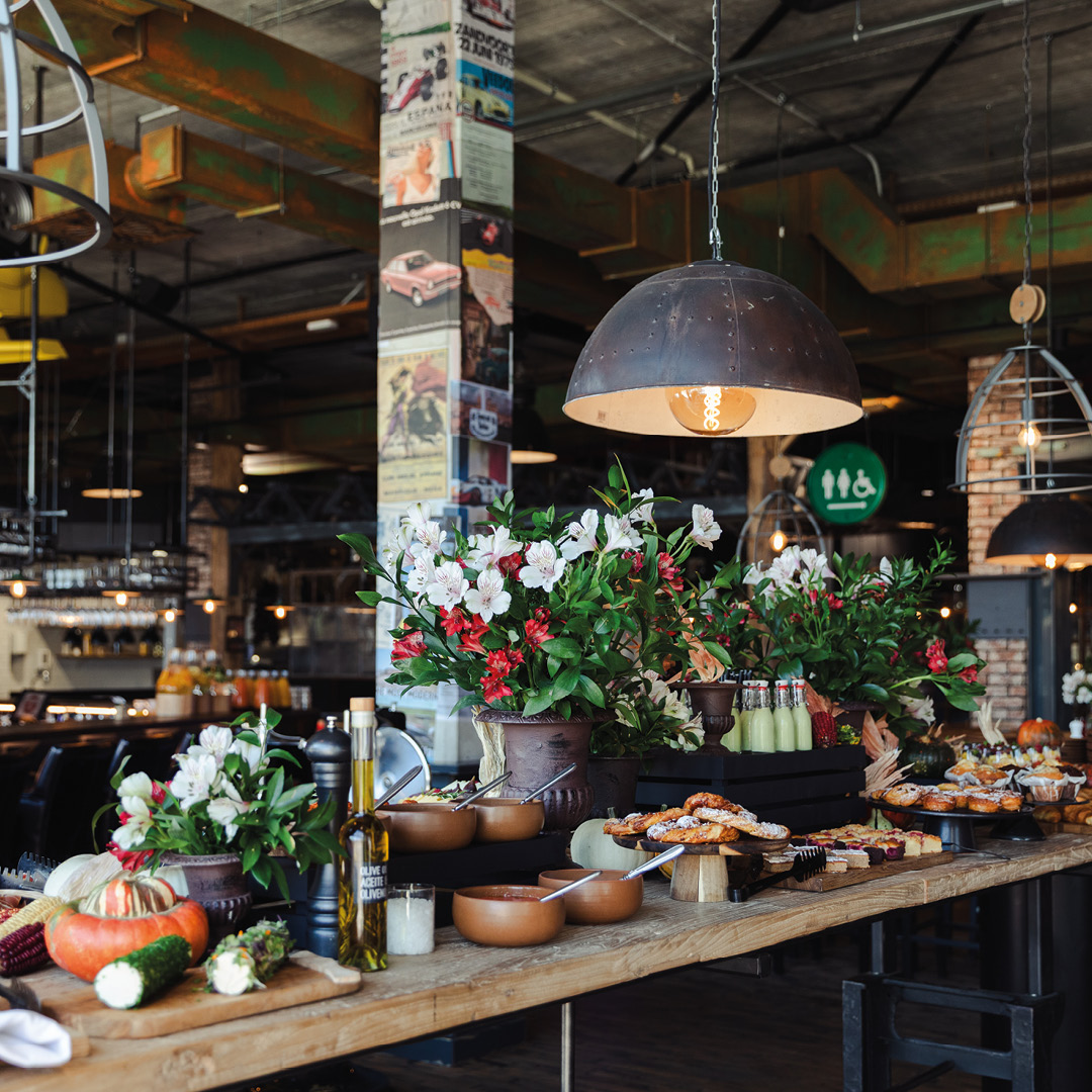 Langer Holztisch in einem Industrie-Café mit Blumenarrangements, Brot, Gebäck und Olivenöl, beleuchtet von einer großen Pendelleuchte.